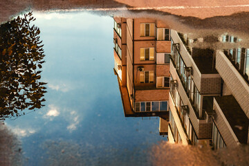 A blurred reflection of an apartment building in the water on the sidewalk of a city street. The...