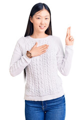 Young beautiful chinese woman wearing casual sweater smiling swearing with hand on chest and...