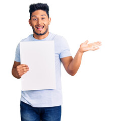 Handsome latin american young man holding cardboard banner with blank space celebrating victory with happy smile and winner expression with raised hands