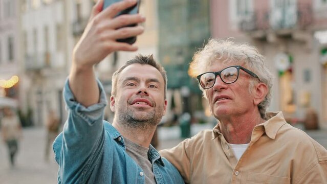 Middle-aged Caucasian male making selfie with older man in middle of street. Men talking to each other. In background people passing by in crowded street. Holding smartphone high for best shot.