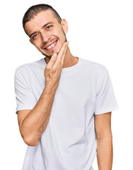 Hispanic young man wearing casual white t shirt looking confident at the camera smiling with crossed arms and hand raised on chin. thinking positive.