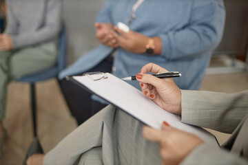 Closeup of unrecognizable female therapist holding clipboard and pen taking notes in support group...