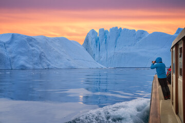 Tourist in a boat in front of an iceberg taking photos at midnight sun sunset. Tourist man explorer watching a large iceberg melting in the arctic sea from the coast of Ilulissat. Greenland - obrazy, fototapety, plakaty