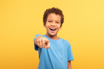 Laughing boy pointing to camera wearing blue t-shirt on yellow background