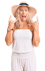 Middle age fit blonde woman wearing summer hat shouting with crazy expression doing rock symbol with hands up. music star. heavy concept.