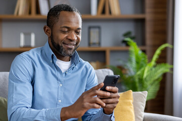 Happy african american man sitting on sofa at home and using mobile phone smiling. Close-up photo. - obrazy, fototapety, plakaty