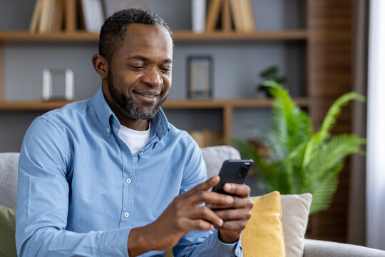 Happy African American Man Sitting On Sofa At Home And Using Mobile Phone Smiling. Close-up Photo.