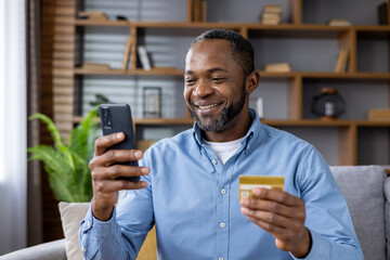 A smiling African American man is at home, sitting on the sofa, holding a credit card and using a...