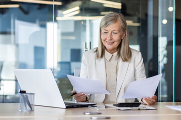 Smiling senior business woman working in office at desk, holding documents in hands, reviewing deal and invoices.