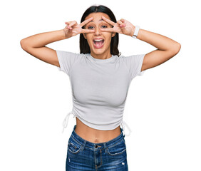 Young hispanic girl wearing casual white t shirt doing peace symbol with fingers over face, smiling cheerful showing victory