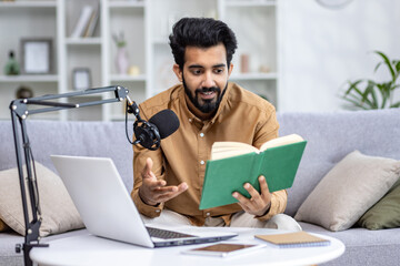 Indian smiling young man sitting on sofa at home in front of laptop and desk with microphone,...