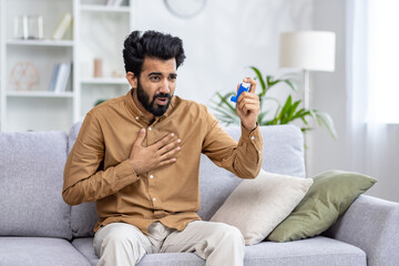 A young Indian man suffers from asthma and allergies. Sitting at home on the couch, holding his...