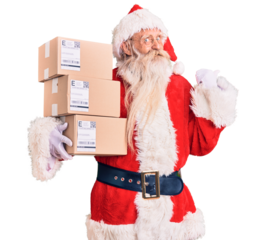 Old senior man with grey hair and long beard wearing santa claus costume holding boxes pointing thumb up to the side smiling happy with open mouth