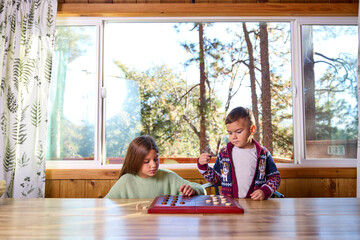 Two siblings play checkers on a table  in a cabin in the woods