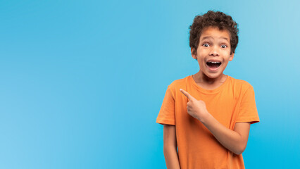 Astonished boy pointing at copy space, excited on blue backdrop