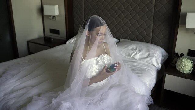 The bride sits on a large bed and holds perfume in her hands