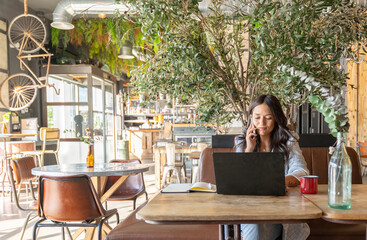 Woman talking on the phone while working online with a laptop computer in a coffee shop.