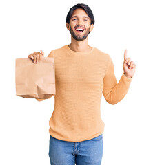 Handsome hispanic man holding take away paper bag surprised with an idea or question pointing finger with happy face, number one