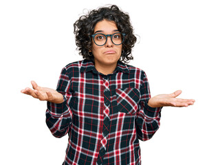 Young hispanic woman with curly hair wearing casual clothes and glasses clueless and confused...