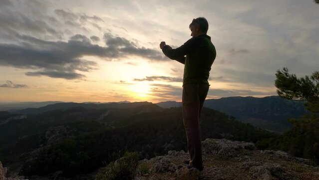 A man in his 40s standing on the top of a mountain records a video of the mountainous landscape at sunset with his smartphone