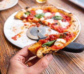 Male hand taking a slice of freshly baked Neapolitan Margherita Pizza on a rustic wooden table in a traditional Pizzeria. - 710929023