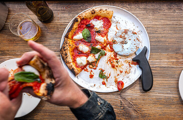 Male hand taking a slice of freshly baked Neapolitan Margherita Pizza on a rustic wooden table in a traditional Pizzeria. - 710929076