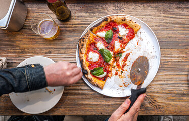 Male hands cutting a freshly baked Neapolitan Margherita Pizza on a rustic wooden table in a traditional Pizzeria. - 710929079