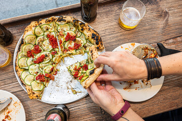 Male hand taking a slice of freshly baked Neapolitan zucchini pizza on a rustic wooden table in a traditional Pizzeria. - 710929240