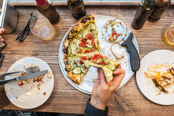 Male hand taking a slice of freshly baked Neapolitan zucchini pizza on a rustic wooden table in a traditional Pizzeria. - 710929259