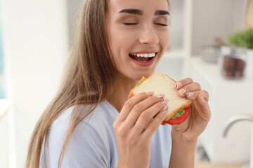 Beautiful young woman eating tasty sandwich in kitchen, closeup