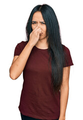 Young hispanic girl wearing casual t shirt smelling something stinky and disgusting, intolerable...