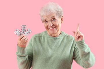 Senior woman with blisters of pills pointing at something on pink background