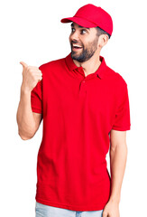 Young handsome man with beard wearing delivery uniform smiling with happy face looking and pointing to the side with thumb up.