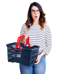 Beautiful young brunette woman holding supermarket shopping basket scared and amazed with open mouth for surprise, disbelief face