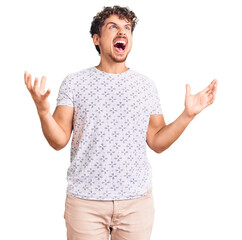 Young handsome man with curly hair wearing casual clothes crazy and mad shouting and yelling with...