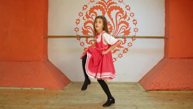 Little girl dressed in red sarafan dances at wooden stage.