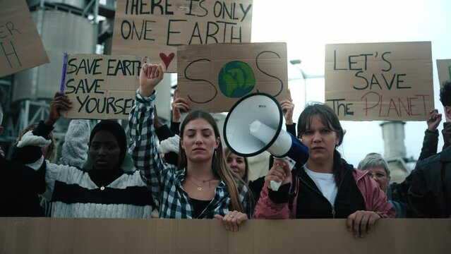 Group of people of different ages and nationalities with arms raised in protest against factories and pollution. Pro-earth demonstration with climate change banners looking at camera with serious face