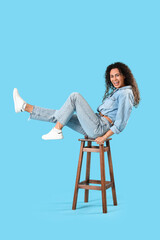Young African-American woman sitting on stool against blue background