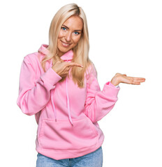 Young blonde woman wearing casual sweatshirt amazed and smiling to the camera while presenting with hand and pointing with finger.