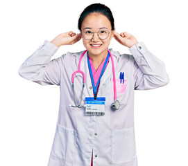 Young chinese woman wearing doctor uniform and stethoscope smiling pulling ears with fingers, funny...
