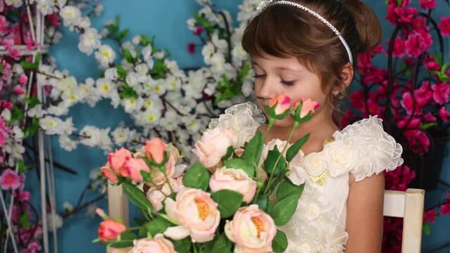 little girl sits on bench among flowers and holds bouquet