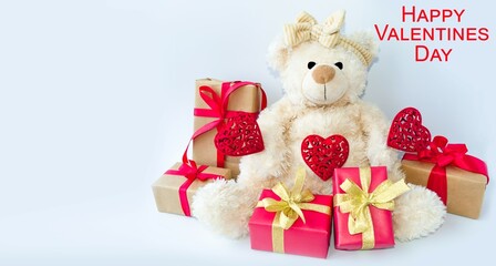 Holiday card for the holiday of Valentine's Day, a toy teddy bear sits on a light background with red hearts and boxes with gifts.  Space for copying text, front view.
