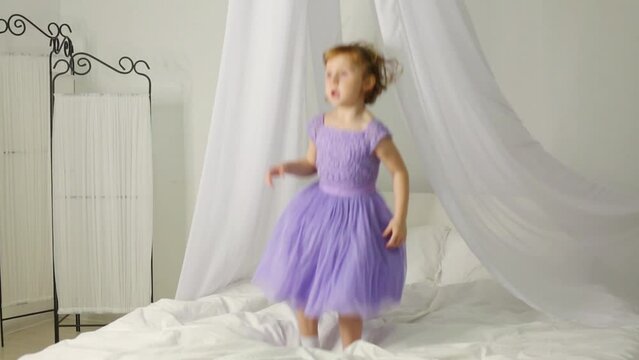 Little girl in dress is jumping on the bed with white baldachin.