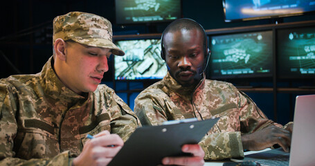 Mixed-races military male workers sitting in monitoring center and studying data from conflict...