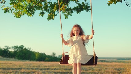 Little girl plays on wooden swing, dreams of flying. Carefree girl swings on swing in park under tree. Baby swing, kid girl smile in flight. Concept of family happiness, dream, Child play in nature.