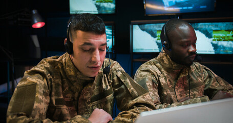 Mixed-races male dispatchers in army co-workers in headsets sitting in military technical room with...