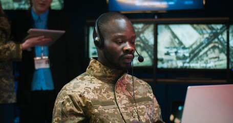 African American military man in headset sitting at laptop computer and talking via videochat in...