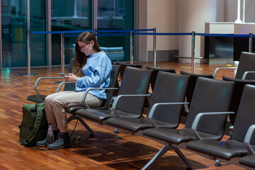 Airport terminal: woman sitting at the airport lounge, waiting for the flight. Thoughtful woman sitting on airport chair using the smartphone.
