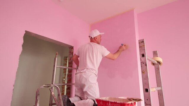 Painter in white clothes works on scaffolding in pink room