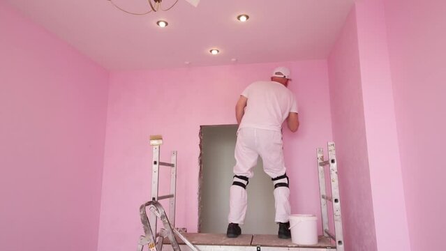 Man in white with brush works on scaffolding in pink room